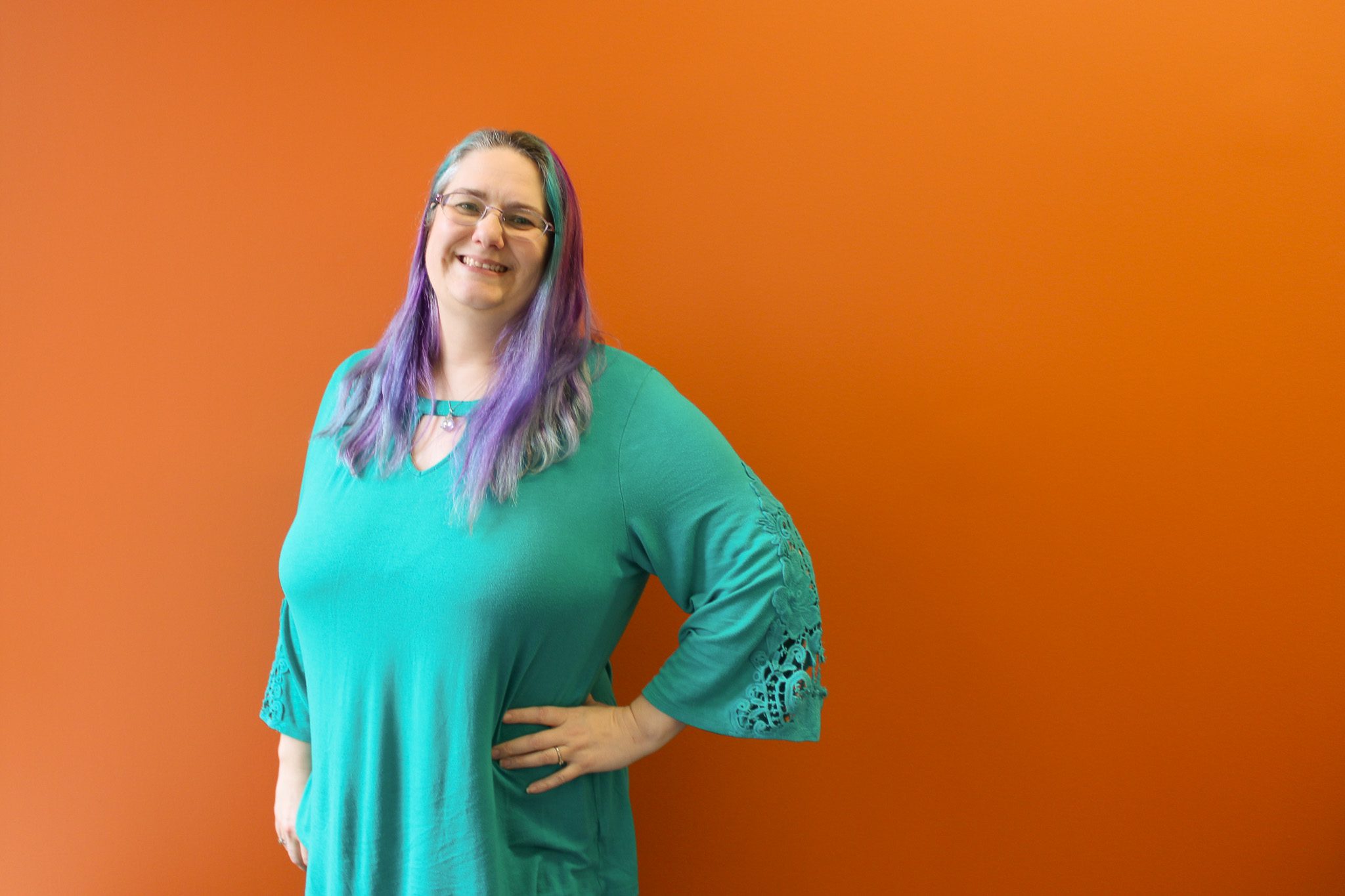 A person wearing a green top stands against an orange background, with their face obscured for privacy. The individual has long, multi-colored hair with prominent shades of blue and purple. They are posing with one hand on their hip.
