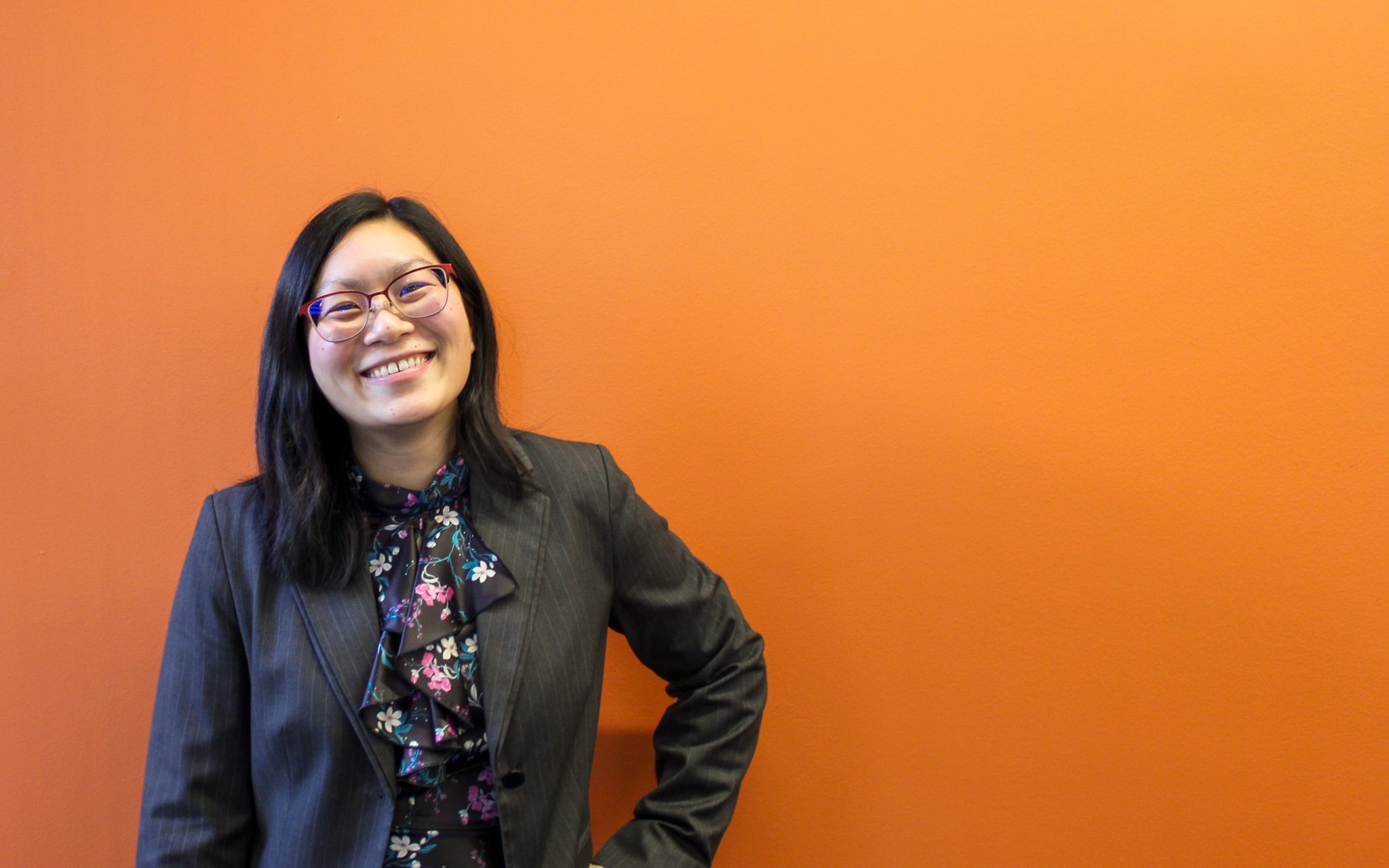 Amy Kuo, a person wearing a grey blazer and a floral dress stands against an orange background. The individual has dark hair that falls to their shoulders, and their posture is relaxed with one hand resting on their hip.