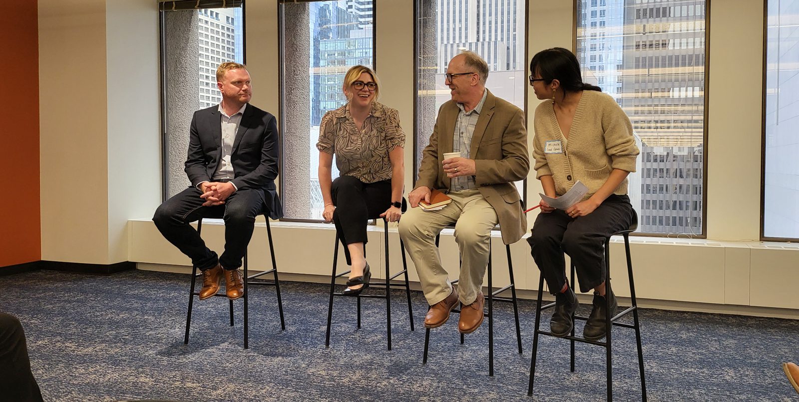 Four individuals are seated on high stools in a panel discussion setup. The individuals from left to right are Blake Webber, Lorraine Glynn, Jim Wolch, and Michelle Chung. The background features large windows with a view of tall buildings.